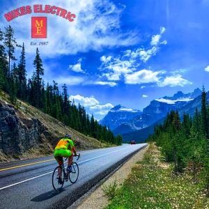 A cyclist rides along the road on the Icefields Parkway in Alberta. The photo is branded with a Mikes Electric Banff logo watermark